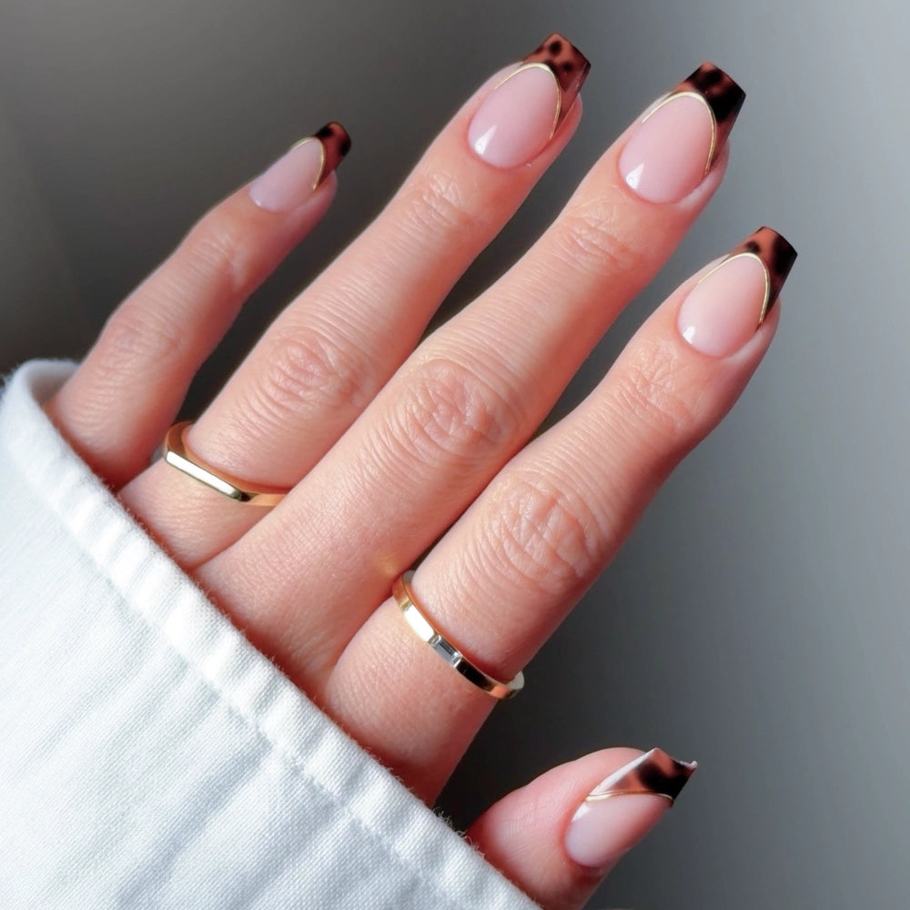 A hand with glamnetic’s Top Tier almond-shaped press-on nails—nude base, tortoiseshell French tips—wears three gold rings and a white long-sleeve garment. - Product image