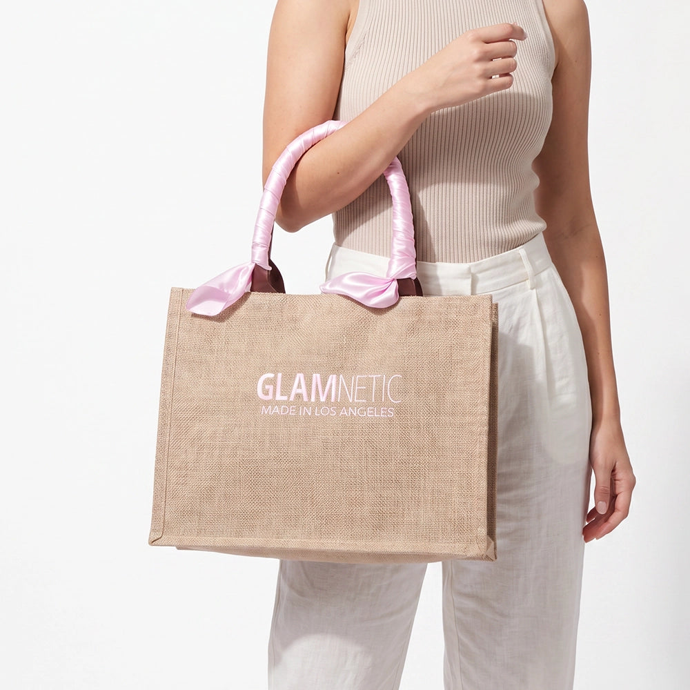 A person in a sleeveless beige top and white pants holds the glamnetic Free Tote Bag ($85+ GWP), a tan jute bag with pink ribbon handles and “GLAMNETIC MADE IN LOS ANGELES” printed on the front. - Product image