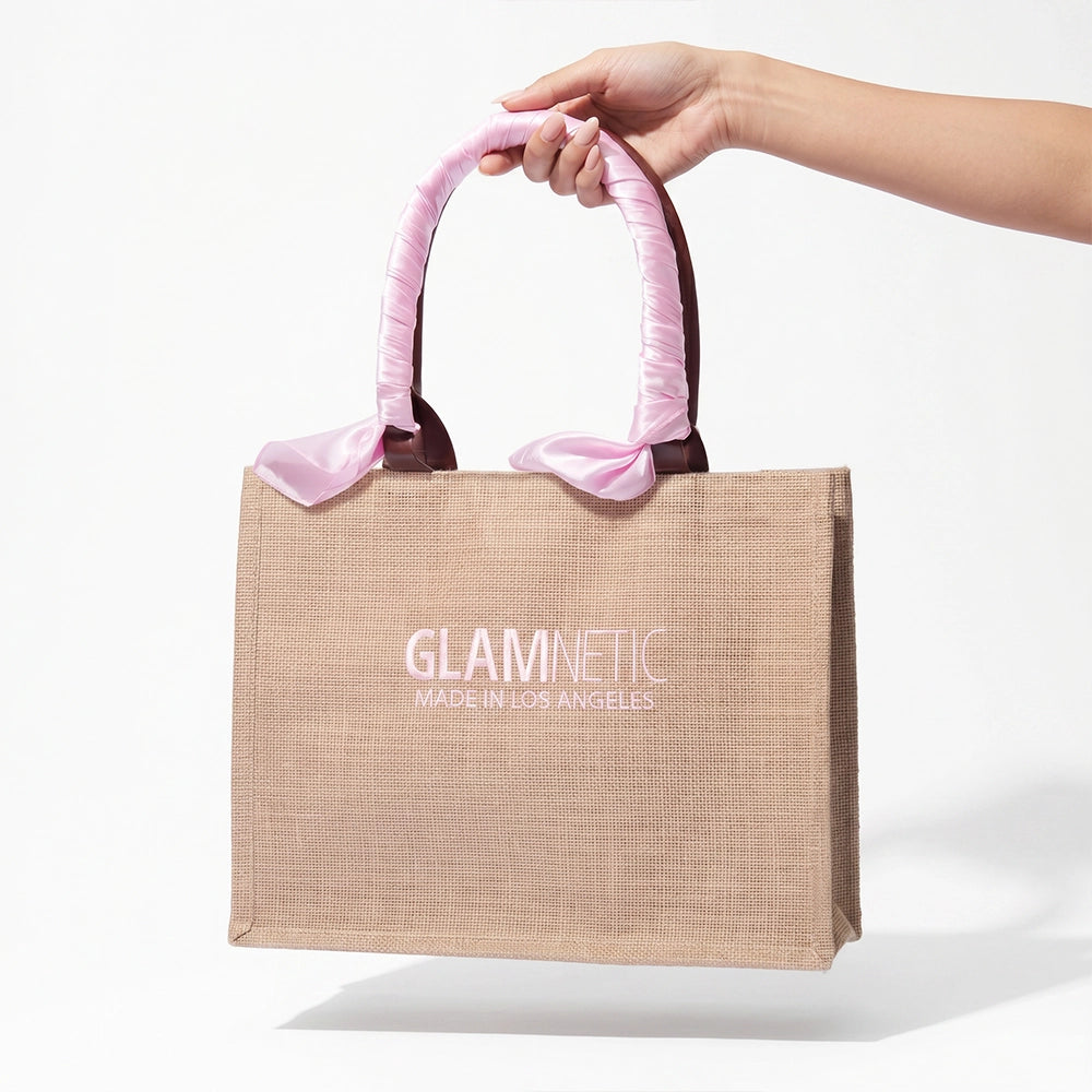 A hand holds the glamnetic Free Tote Bag ($85+ GWP), featuring pink satin-wrapped handles and "GLAMNETIC MADE IN LOS ANGELES" on beige fabric, shown against a plain white background. - Product image