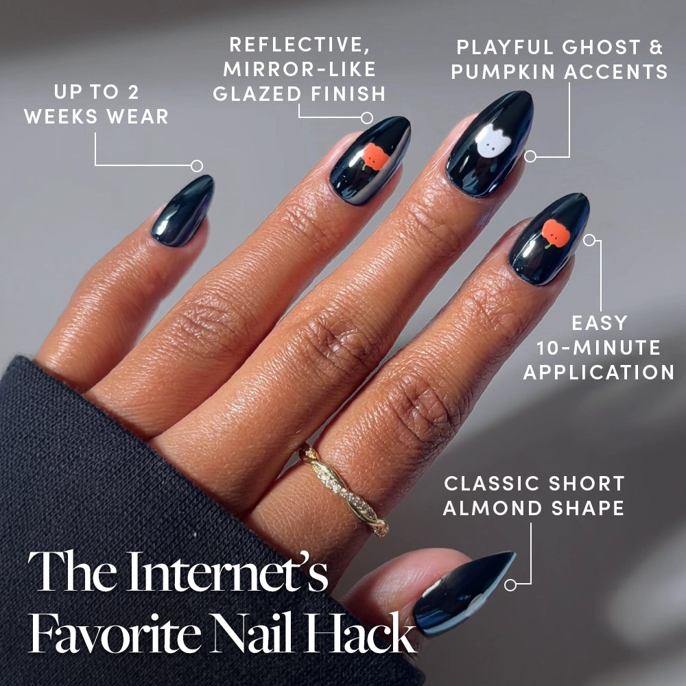 A hand displays short, almond-shaped nails from the glamnetic Tricks & Treats Press-On Nail Set, featuring glossy black polish with white ghost and orange pumpkin accents for a festive Halloween look. The set boasts up to 2 weeks of wear, an easy 10-minute application, and a glazed finish that delivers a striking, reflective shine. - Product image
