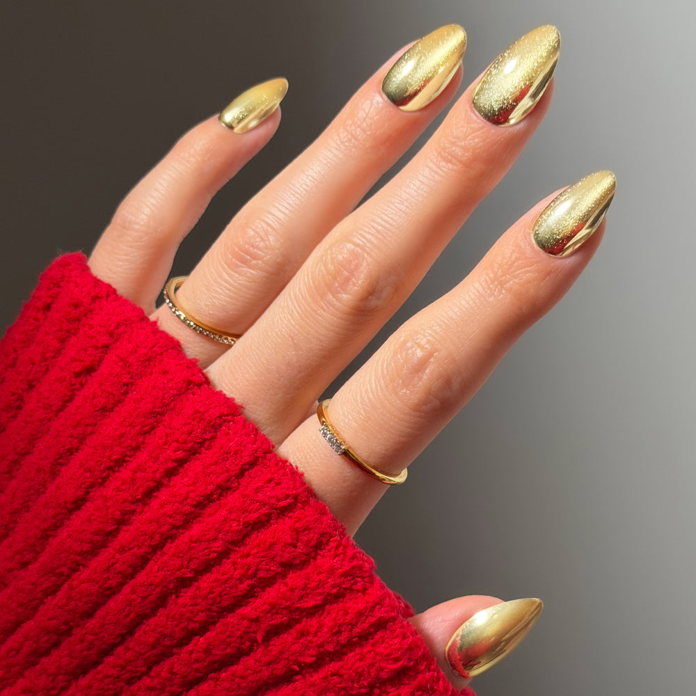A hand with short almond gold chrome Trophy Wives nails wearing two thin gold rings against a red knit sleeve.