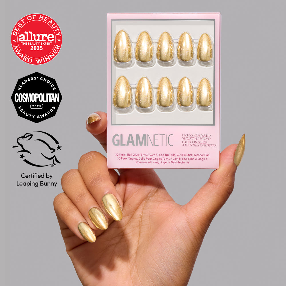 A hand with short, metallic gold almond press-on nails holds a boxed set of Glamnetic Trophy Wives, with Allure and Cosmopolitan award seals and a Leaping Bunny certification logo displayed nearby. - Product image