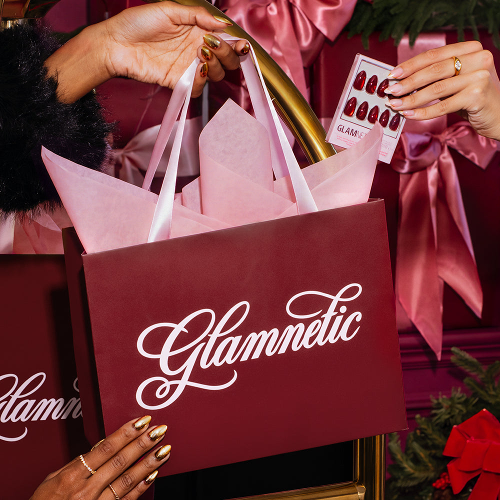Hands holding a maroon shopping bag with a white logo and a package of glossy red short almond Trophy Wives press-on nails. - Product image