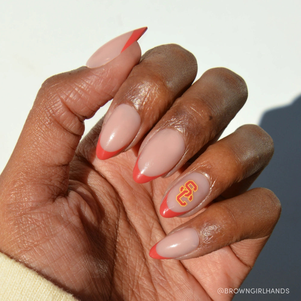 A hand with medium brown skin wears glamnetic's USC Trojans Press-On Nail Set, showing almond-shaped nails with a nude base, red French tips, and one nail featuring the yellow and red USC logo against a plain light background. - Product image