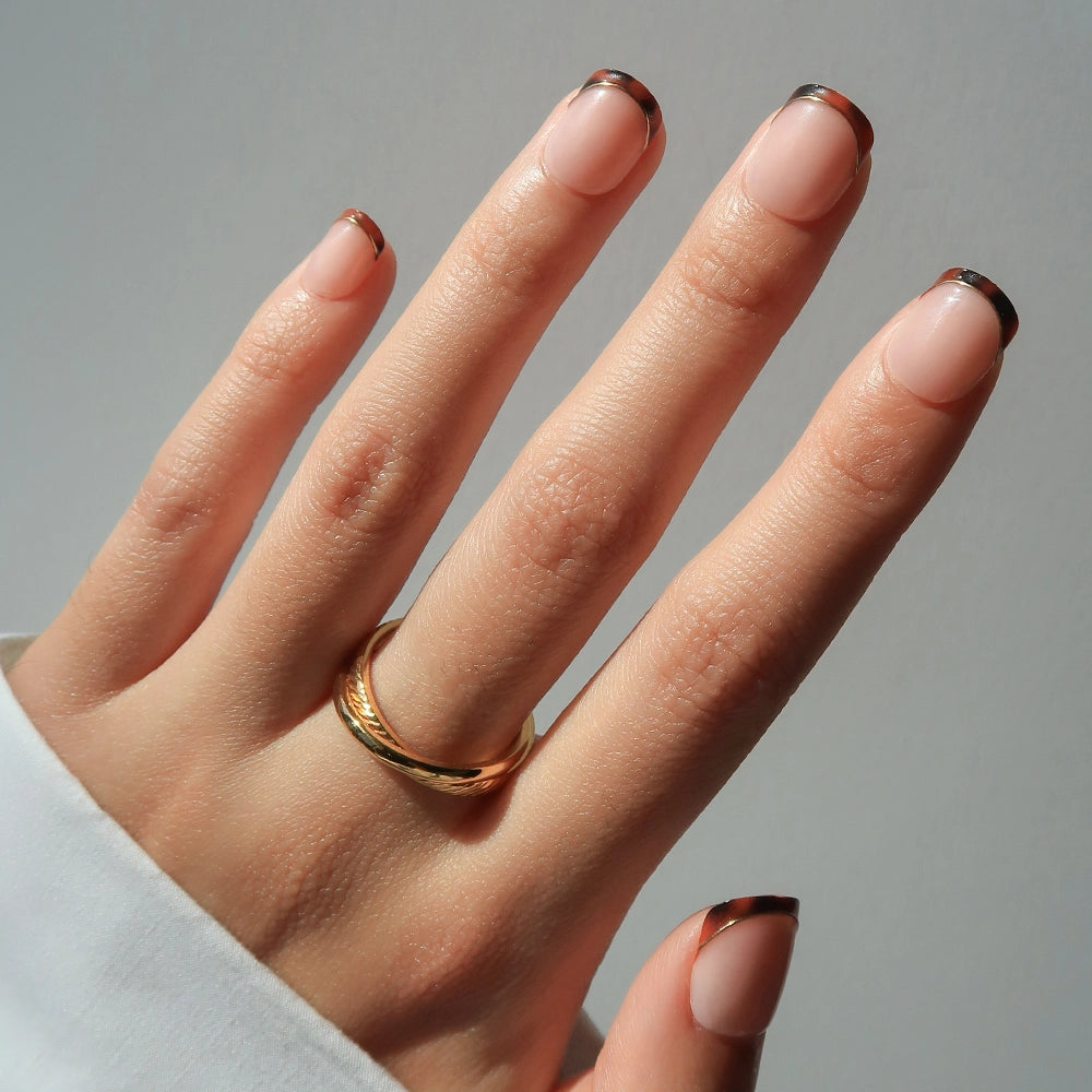 A hand adorned with a gold ring wears glamnetic’s Unbothered press-on nails—super short, nude base with dark brown tortoiseshell French tips—posed against a light background with a white long-sleeve shirt.