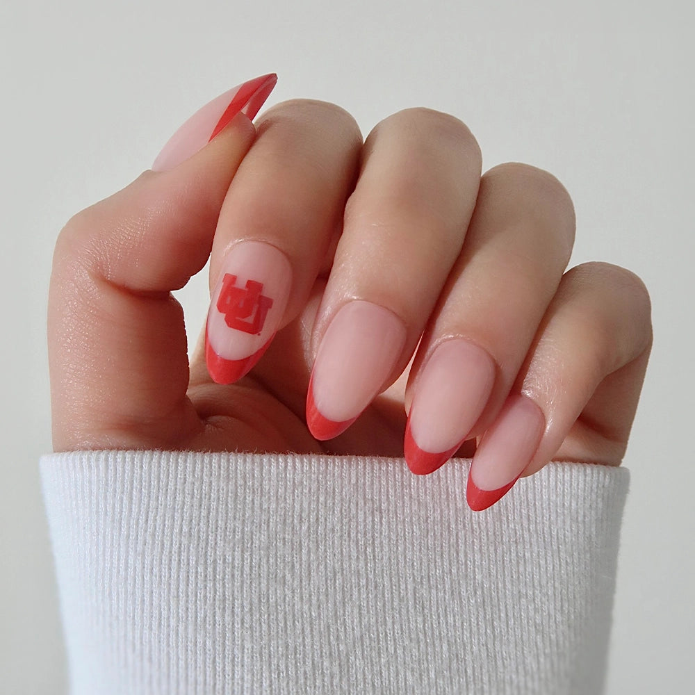 A hand wears the glamnetic Utah Utes Press-On Nail Set, featuring salon-quality almond nails with red French tips and a bold Utah Utes logo on the ring finger. The person is dressed in a white long-sleeve top. - Product image