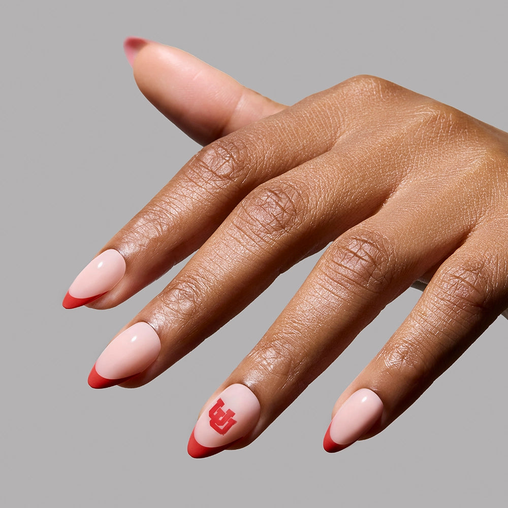 A hand with almond-shaped, pale pink nails featuring red French tips from the glamnetic Utah Utes Press-On Nail Set. A red graphic on the ring finger pops against a solid gray background, highlighting these salon-quality press-ons. - Product image