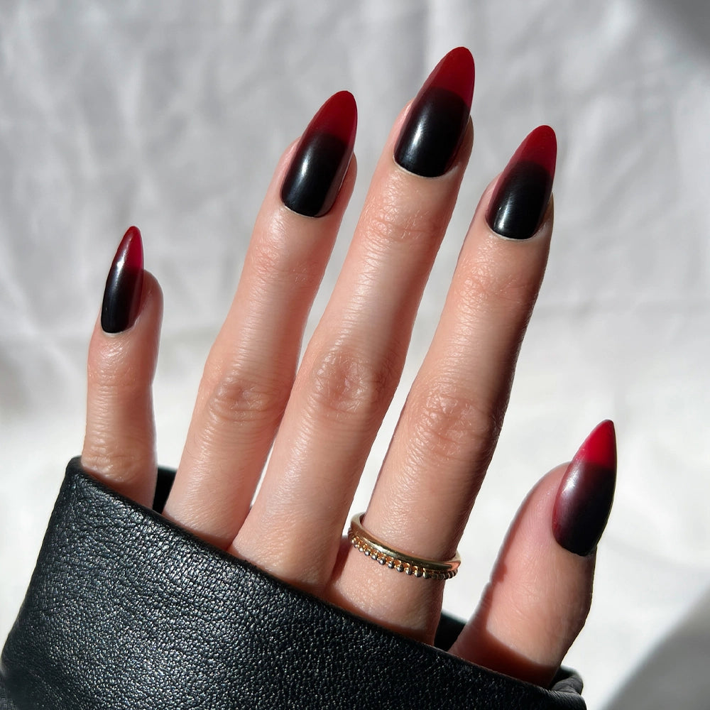 A hand with long, almond-shaped nails showcases the striking black-to-red ombre design from the glamnetic Vampiress press-on nail set. The person wears a gold ring and a black jacket, fingers slightly spread against a light background—a salon-quality mani perfect for anyone seeking bold almond press-on nails.