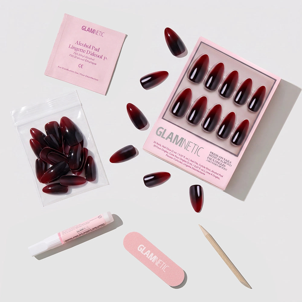 Get a salon-quality manicure at home with the glamnetic Vampiress Press-On Nail Set. This set includes dark red almond-shaped press-on nails, nail glue, a mini nail file, a wooden cuticle stick, extra nails in a bag, and an alcohol pad—all elegantly displayed on a light surface. - Product image