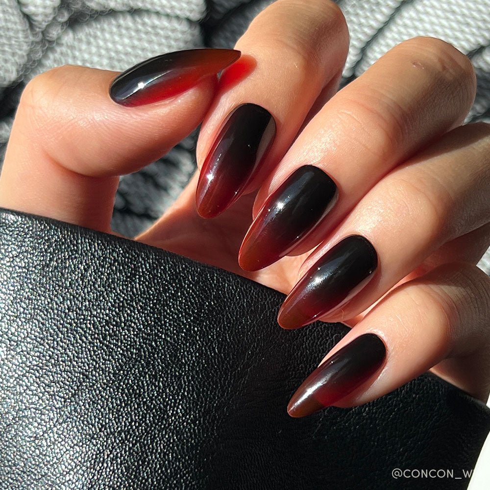 A hand with long, almond-shaped nails featuring a glossy black-to-blood ombre design from glamnetic's Vampiress set, holding a textured black object in bright sunlight.
