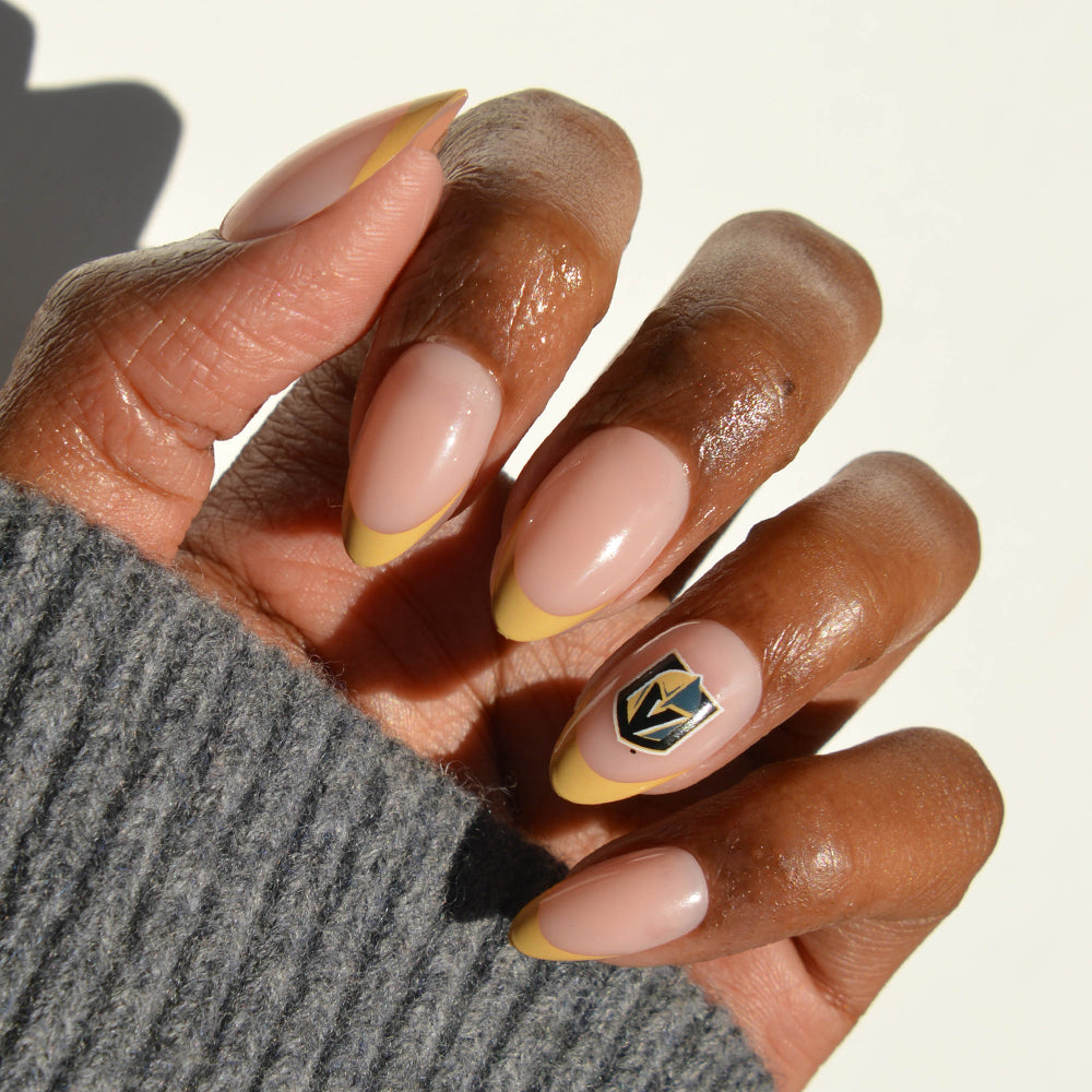 A hand wearing glamnetic NHL Press-On Nails showcases long, almond-shaped tips in nude with yellow accents; the ring fingernail features a striking black and gold shield design. The person is dressed in a gray sweater. - Product image