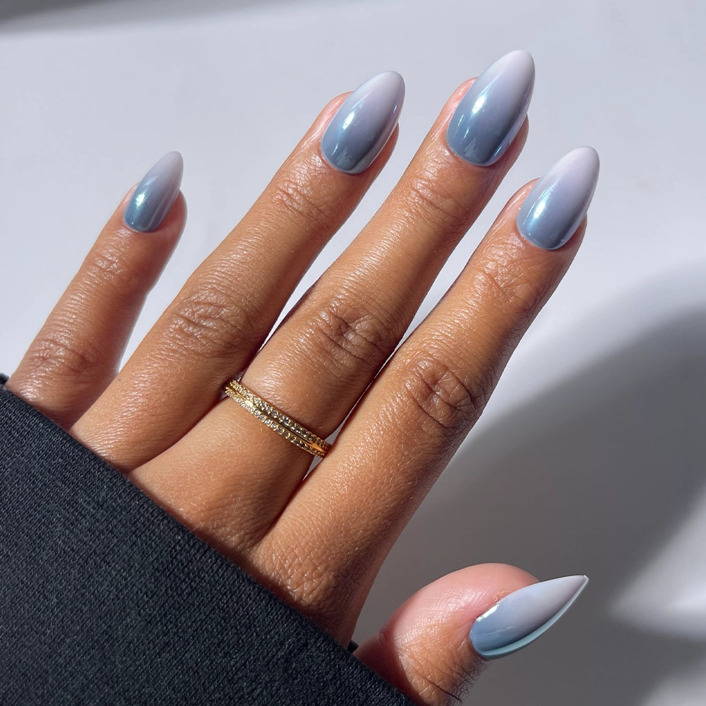 A hand with long, almond-shaped nails painted in the Vintage Silver design by glamnetic, featuring a blue-to-white ombre reminiscent of a glazed nail set. The person wears a gold ring and has medium brown skin, with a black sleeve. The background is white with soft shadows.