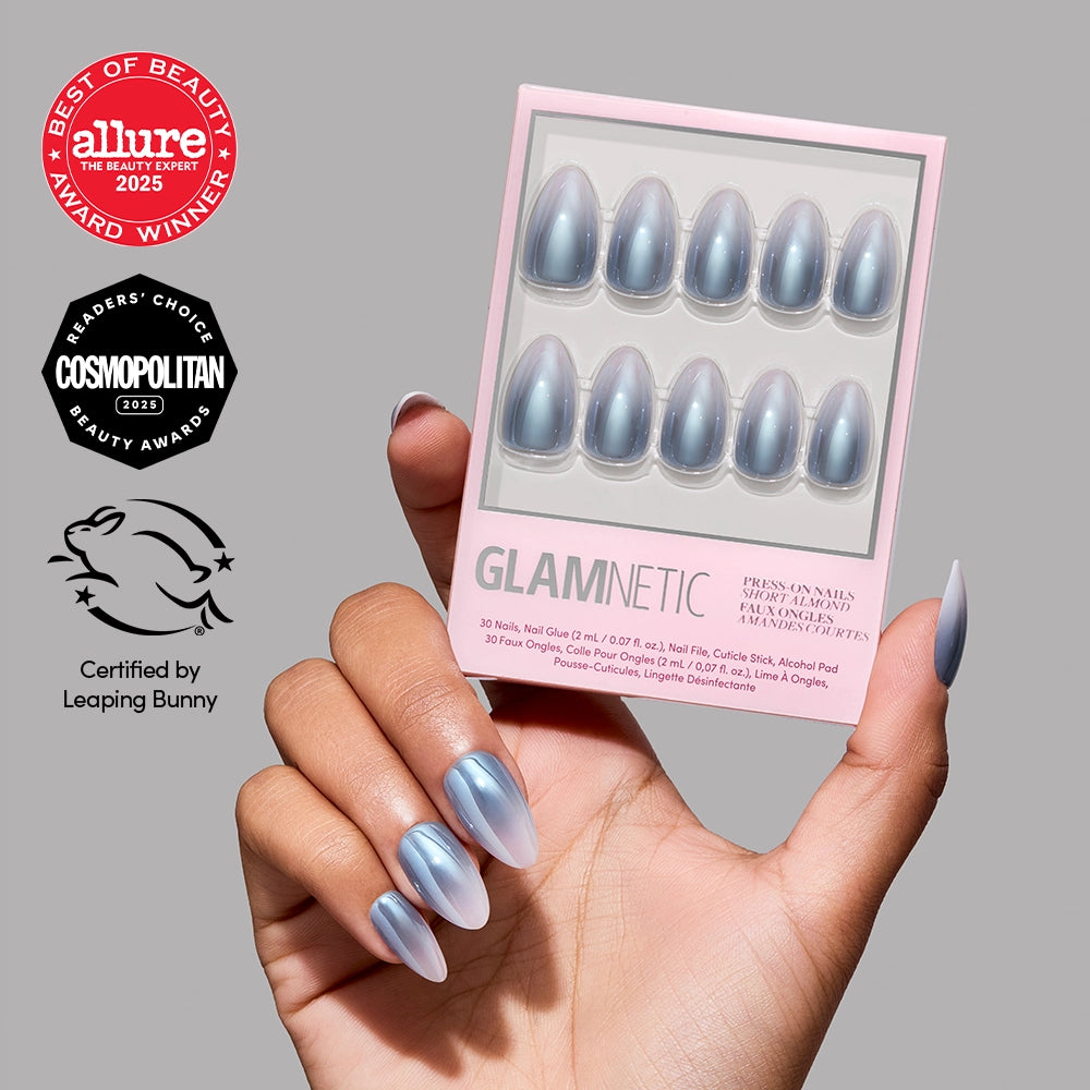 A hand holds a pink box of glamnetic Vintage Silver short almond nails. The shimmering blue-grey ombre nails evoke a classic press-on style. The image features Allure and Cosmopolitan 2025 award logos, as well as the Leaping Bunny cruelty-free certification icon, all set against a light grey background.