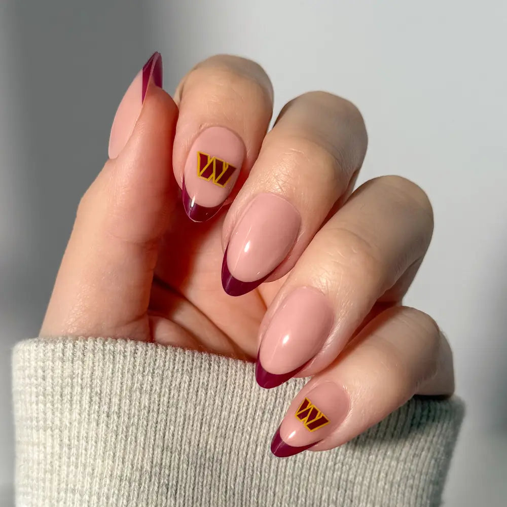 A hand showcases the glamnetic NFL Press-On Nails, styled with almond-shaped nails featuring pink polish, dark purple French tips, and gold “W” decals on the middle and ring fingers. The person is also wearing a light gray ribbed sweater. - Product image