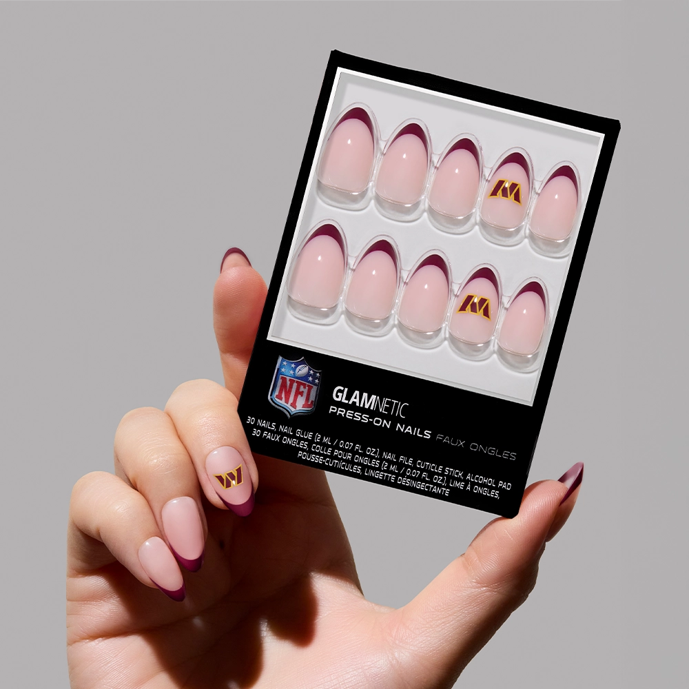 A hand displays the glamnetic Washington Commanders Press-On Nail Set, featuring a pink base, dark red tips, and gold NFL logos on two nails, against a plain light gray background for ultimate fan nail art.