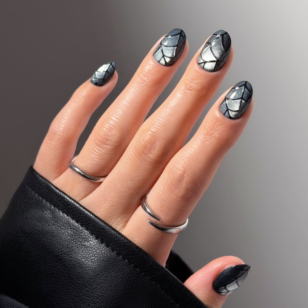 A hand with short oval nails featuring “Weblings” silver and black geometric nail art by glamnetic, accessorized with two silver rings and a black leather sleeve, displayed against a neutral background.