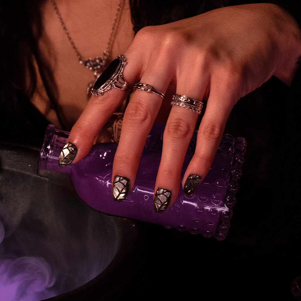 A hand with Weblings black velvet nails by glamnetic, adorned with silver rings, pours a purple potion from a decorative bottle into a smoky cauldron. - Product image