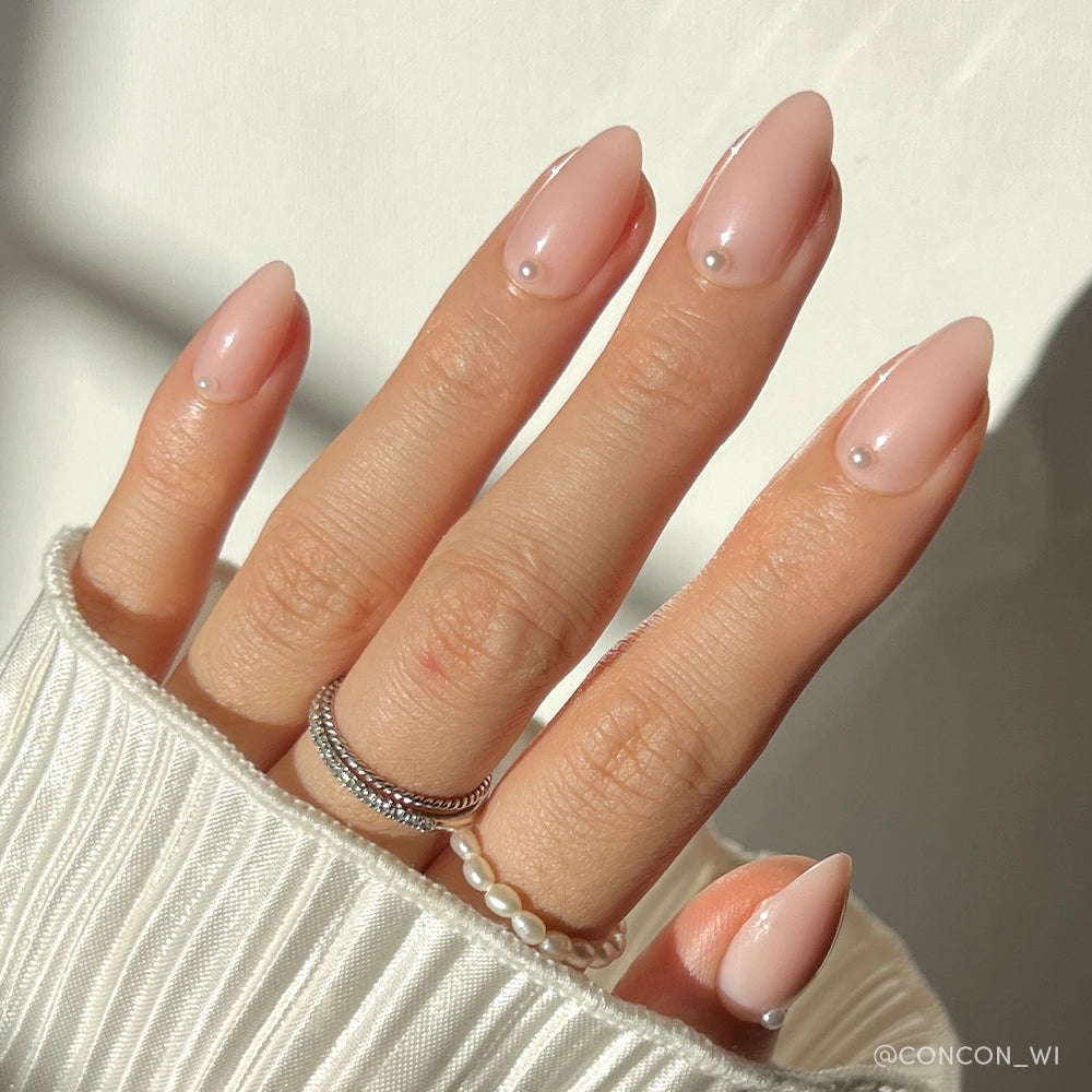 A hand with almond-shaped, nude pink nails from glamnetic’s Wifey set features 3D pearls near each cuticle. Silver and pearl rings plus a ribbed white sleeve enhance the look, as sunlight highlights the salon-quality manicure.