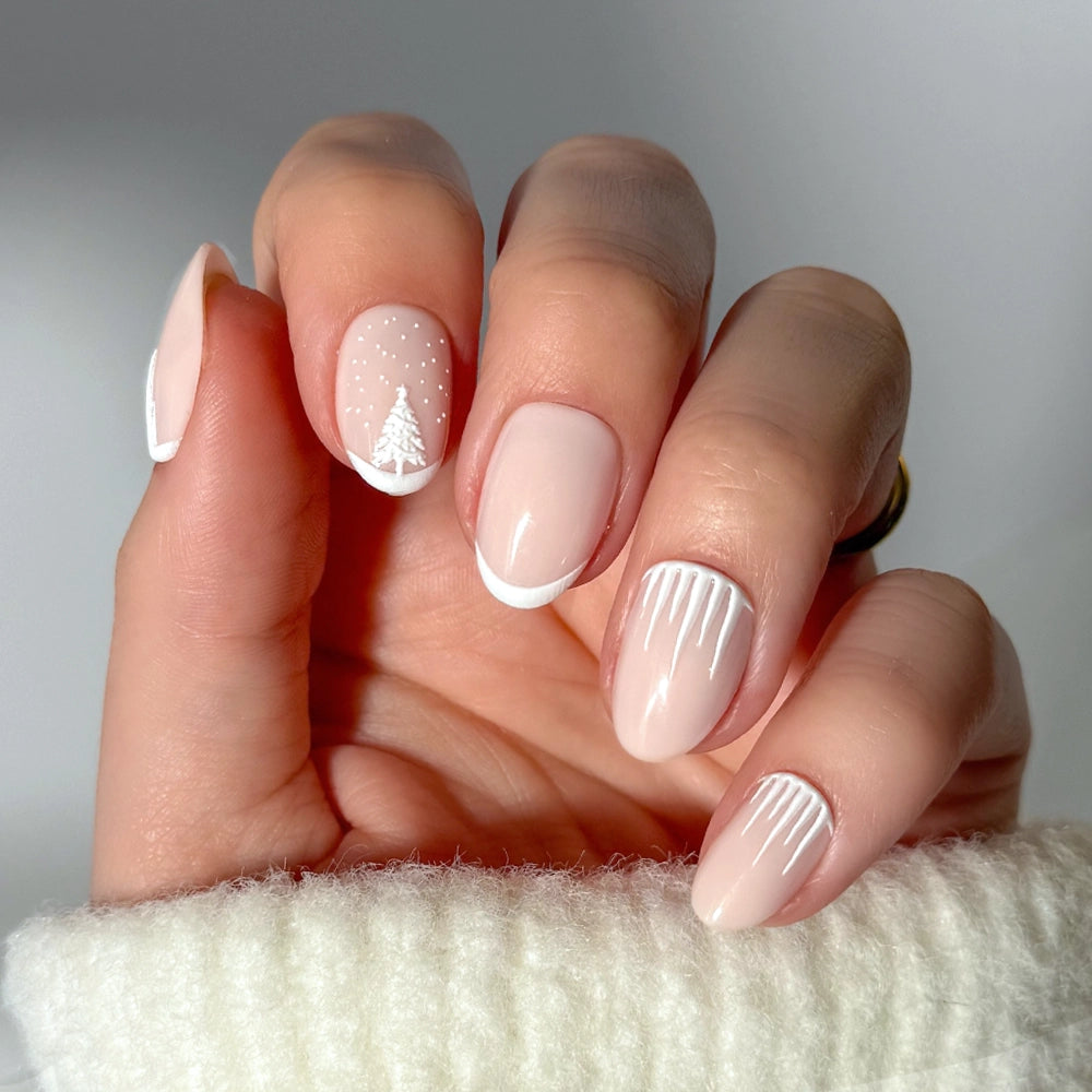 A hand adorned with glamnetic’s “Winter’s Tale” press-on nails—nude and white winter-themed nail art featuring Christmas tree accents, French tips, and icicle designs on short oval nails—is displayed against a cozy white sweater background.