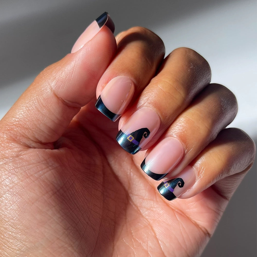 A hand adorned with the Witchy Woman press-on nails by glamnetic features black French tip short squoval nails, accented with small witch hat designs on the ring and middle fingers, all beautifully showcased in natural light for a modern and enchanting manicure.