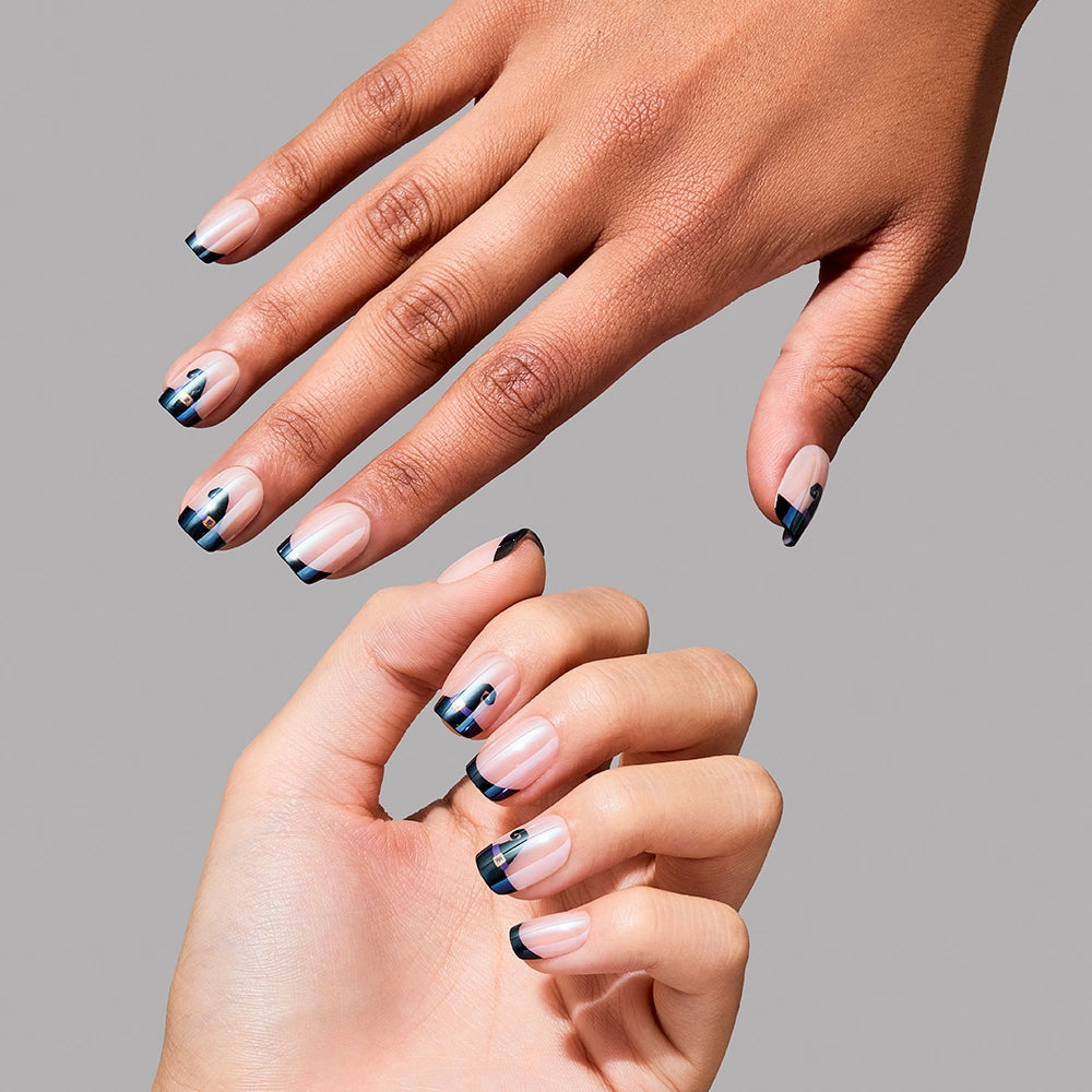 Two hands with manicured nails model the glamnetic Witchy Woman set. These short squoval press-ons display a modern French tip, adorned with navy and teal geometric lines and dots over a natural base, all posed gracefully against a simple gray backdrop. Perfect for anyone wanting stylish, contemporary press-on nails from glamnetic. - Product image
