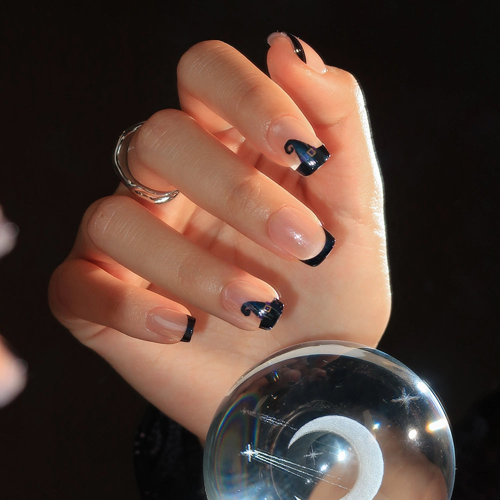 A hand adorned with glamnetic’s Witchy Woman press-on set—featuring long, square-shaped nails with glossy black French tips and abstract swirl accents—holds a clear glass sphere against a dark backdrop. - Product image