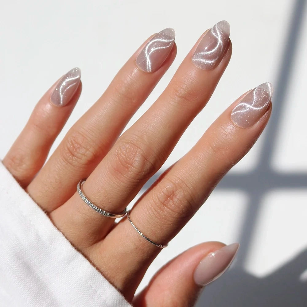A hand with almond-shaped nails featuring WOO-MAN! by glamnetic—sheer, shimmery white press-ons with elegant white swirl designs; the person wears two thin silver rings and a white long-sleeve shirt. These glamorous WOO-MAN! almond press-on nails by glamnetic deliver a flawless, salon-quality look in minutes.
