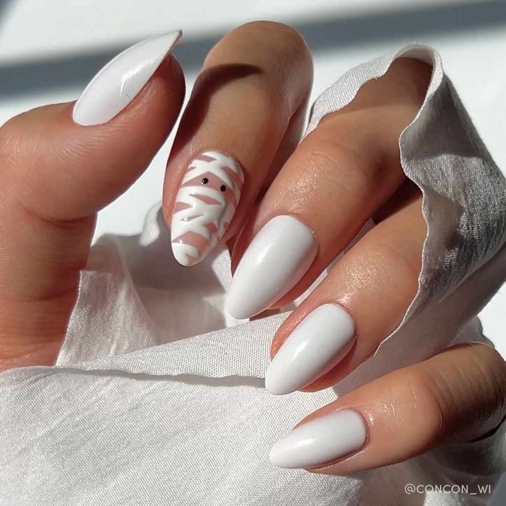 A hand with long, almond-shaped white nails—one nail features a mummy design. The hand holds white fabric, showcasing glamnetics playful Wrapped Up press-on nail set.