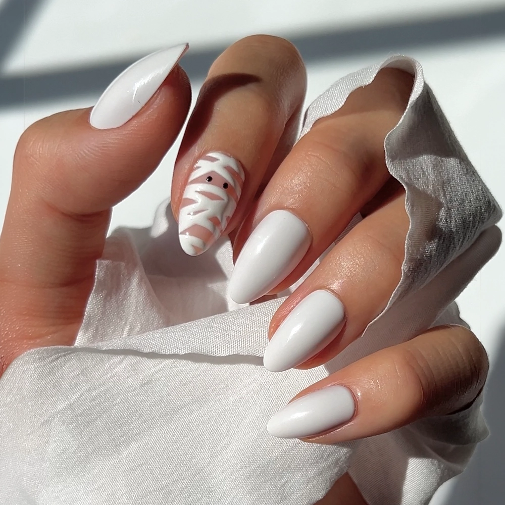 A hand with long, almond-shaped glamnetic “Wrapped Up” press-on nails painted white; the ring fingernail showcases a white mummy nail design with delicate wraps and two small black eyes. The hand holds white fabric in soft, natural light.
