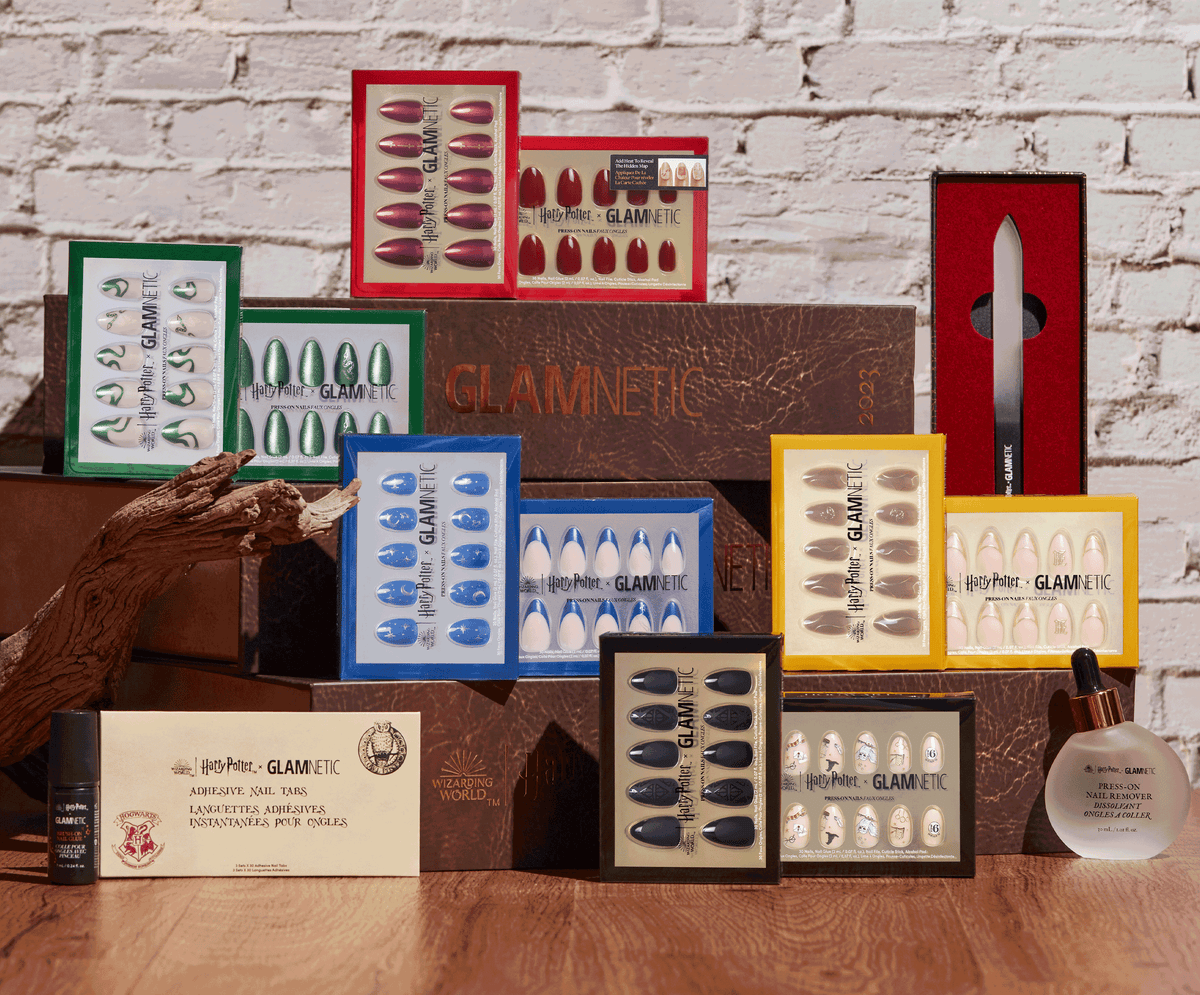 A display of GLAMNETIC press-on nails in various colors and Harry Potter-themed designs, with a nail file, serum bottle, and ornate wand on a wooden surface against a white brick wall.