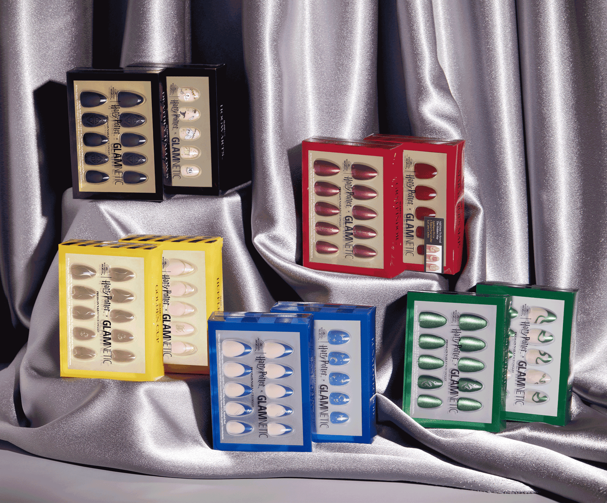 Several boxed sets of press-on nails in different colors and packaging, arranged on shiny silver fabric. The boxes are in red, yellow, blue, green, and black, showing rows of various colored nails inside.