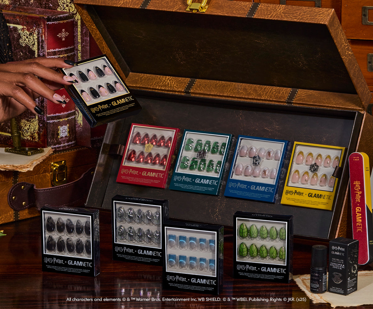 Display of Glamnetic x Harry Potter press-on nails collection arranged in an open vintage trunk, showcasing designs inspired by Hogwarts houses in red, green, blue, and yellow packaging, with themed accessories and props in the background