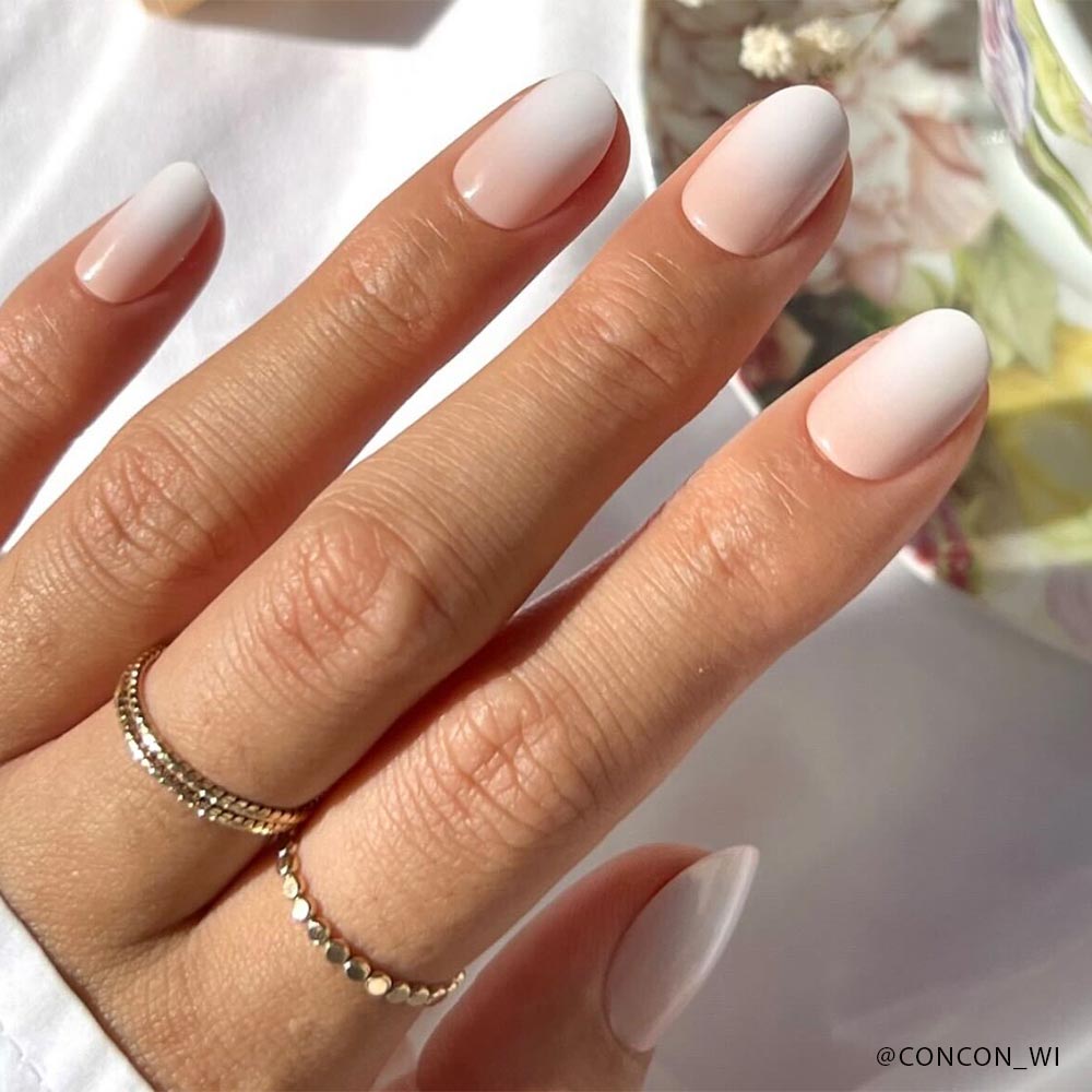 A hand with glamnetic Nail Lover's Gift Set press-on nails in a pink-to-white ombre, paired with delicate gold rings. A softly blurred floral background adds an elegant touch. - Product image