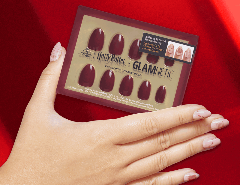 A hand with light pink, patterned nails holds a box of Harry Potter x Glamnetic press-on nails in a deep red color, set against a red fabric background.