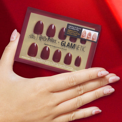 A hand with pale pink nail art holds a box of Harry Potter x Glamnetic press-on nails in a deep red shade, displayed against a rich red fabric background.