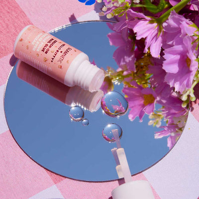 Open bottle of Glamnetic x Hello Kitty Brush-On Nail Glue placed on a round mirror with pink flowers reflected on its surface, styled over a pink checkered background.