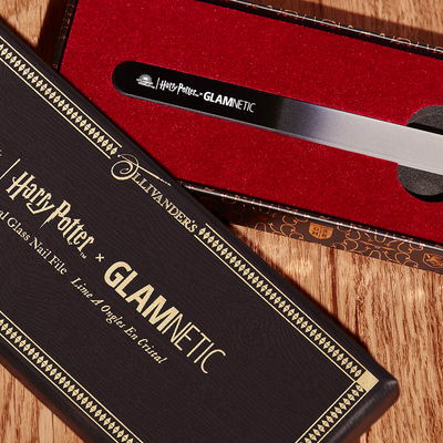 A Harry Potter & Glamnetic nail file in a black and gold box with red velvet lining, displayed on a wooden surface. The file has a black handle with white text and is presented in an open case.