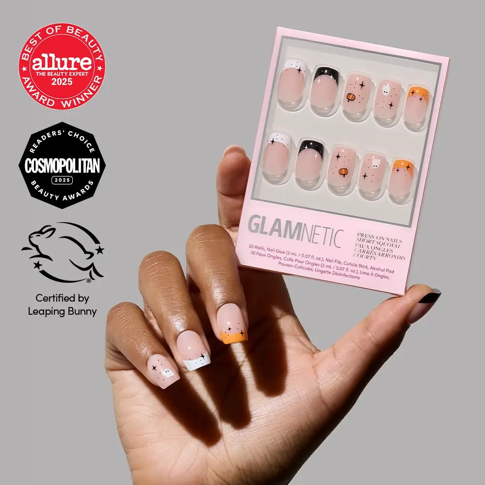 A hand displays black and orange Halloween nails from glamnetic’s Tiny Pumpkin set. The short press-on nails are arranged next to the packaging and three beauty award logos, all set against a plain gray background.