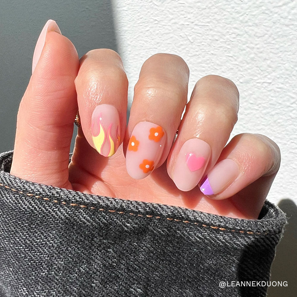 A denim-sleeved hand shows off glamnetics Wild Child nails—short oval, pastel pink with a semi-transparent base and playful designs: yellow and pink flames, orange flowers, a pink heart, and a purple tip shining in sunlight.