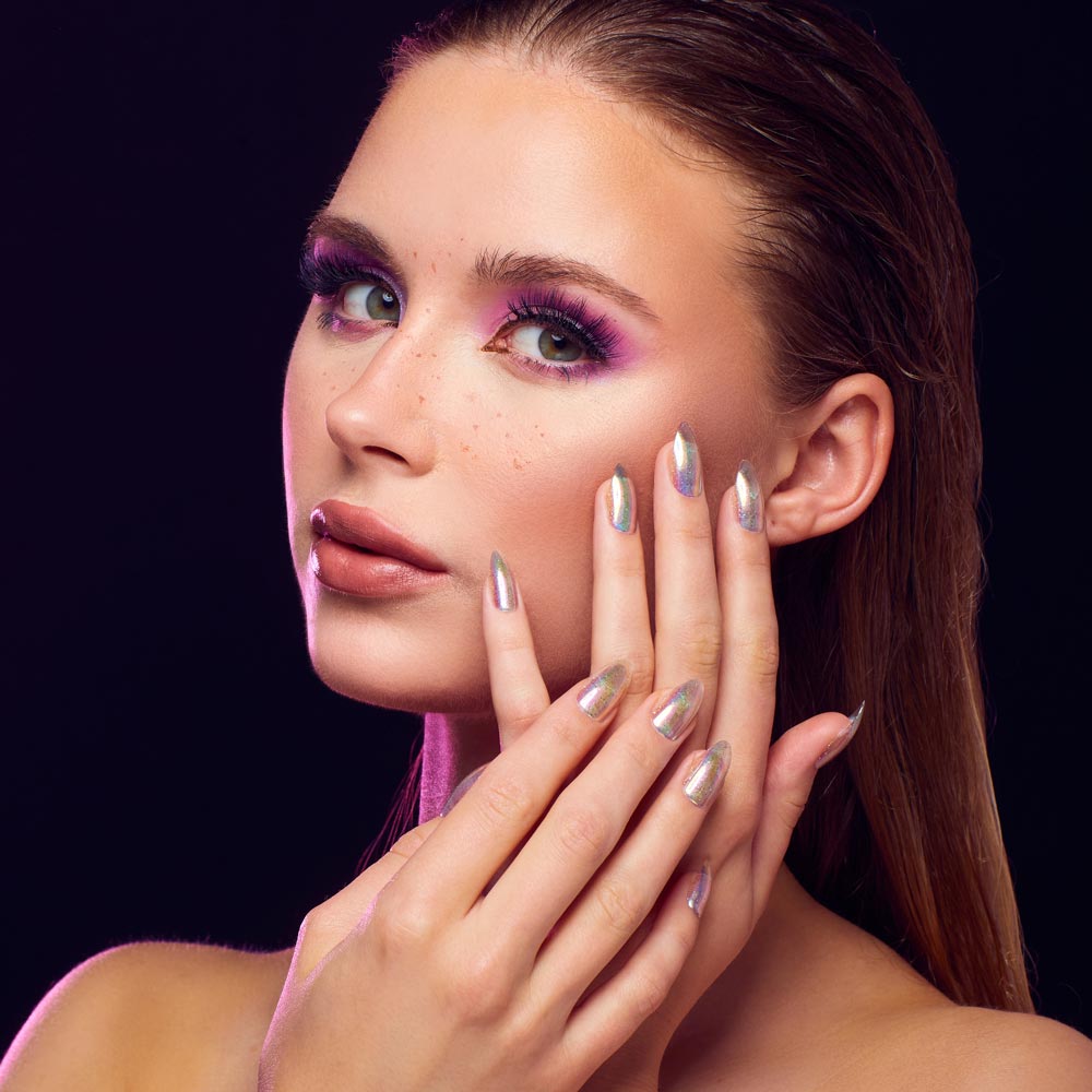 A woman with slicked-back hair showcases glamnetics Stardust pointed almond holographic nails and bold purple eye makeup, touching her face to highlight her sparkling manicure and striking look against a dark background. - Product image