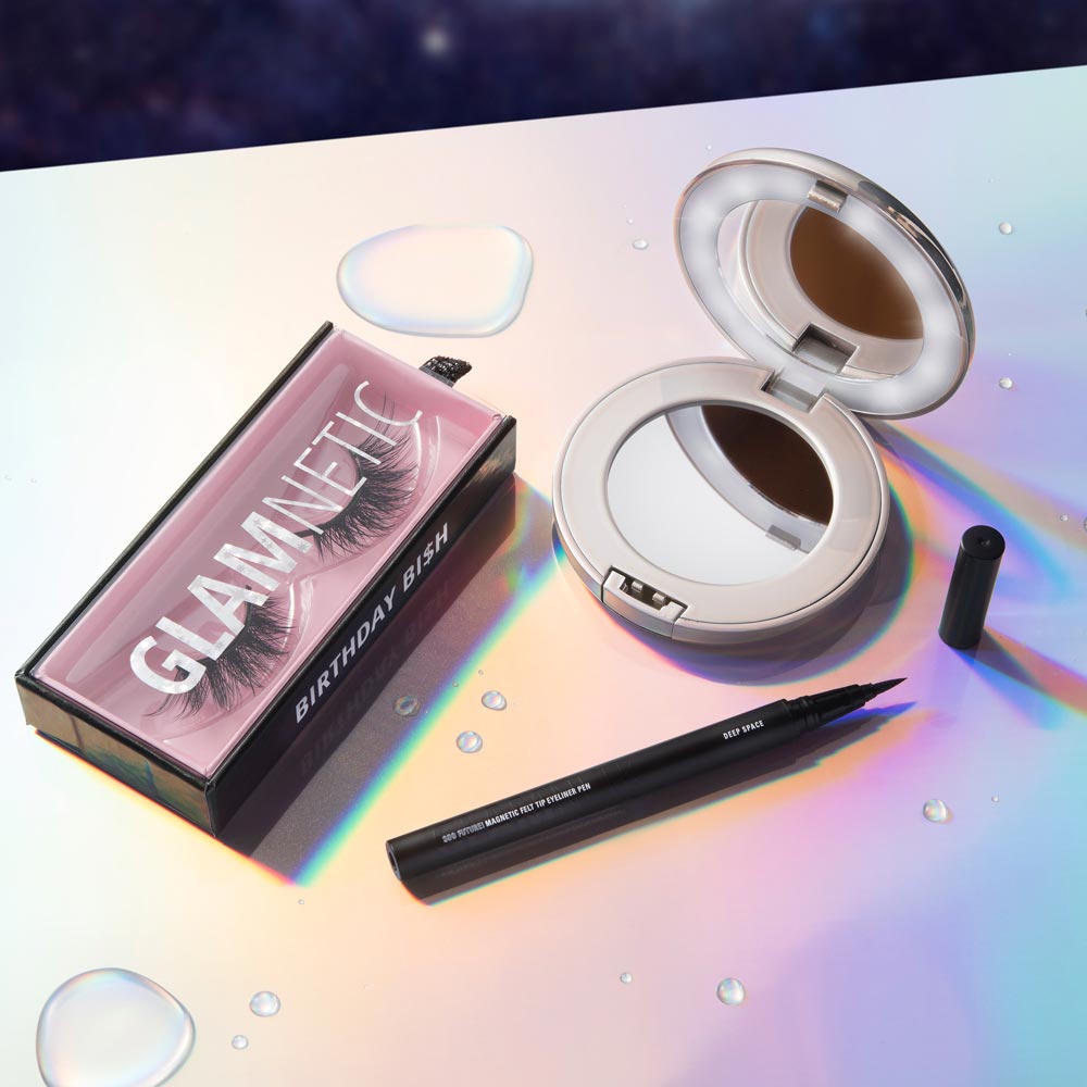 On a shiny surface with water droplets: Glamnetic false eyelashes in a box, the Glamnetic Compact Mirror (with built-in magnification), and an open black liquid eyeliner pen. - Product image