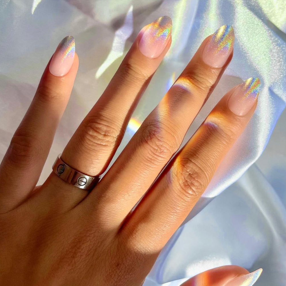 A hand with almond-shaped nails painted in glamnetics Winter Storm holographic polish rests on white satin, reflecting rainbow hues. A simple metallic ring adorns one finger.