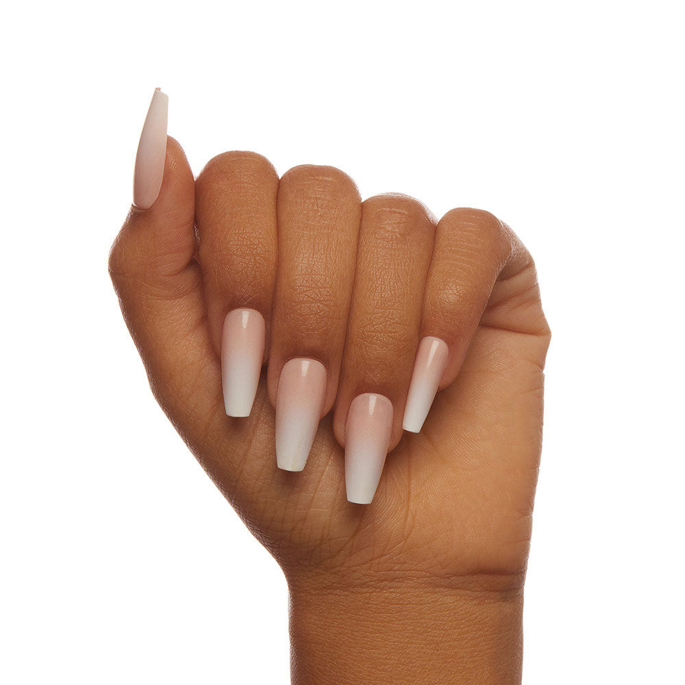 A hand with medium brown skin displays long, coffin-shaped, customizable glamnetic Creme de Nude nails, featuring a glossy light pink to white ombre design, shown in a loose fist against a white background. - Product image
