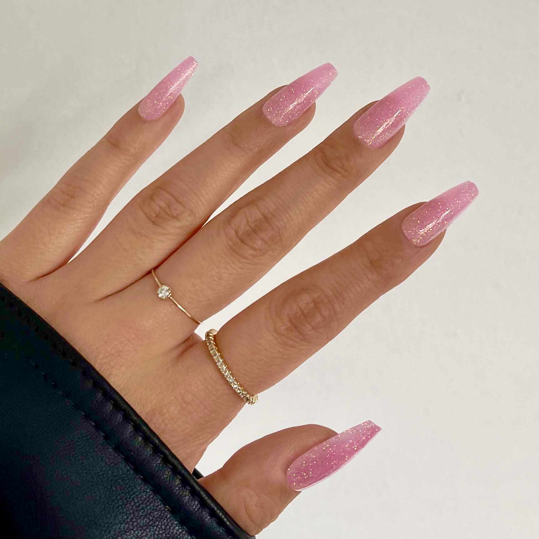 A hand shows off Juicy by glamnetic: long, glossy pink coffin nails with glitter. Two delicate rings—a small stone and a plain band—add charm, while a black sleeve hints at early 2000s style. - Product image