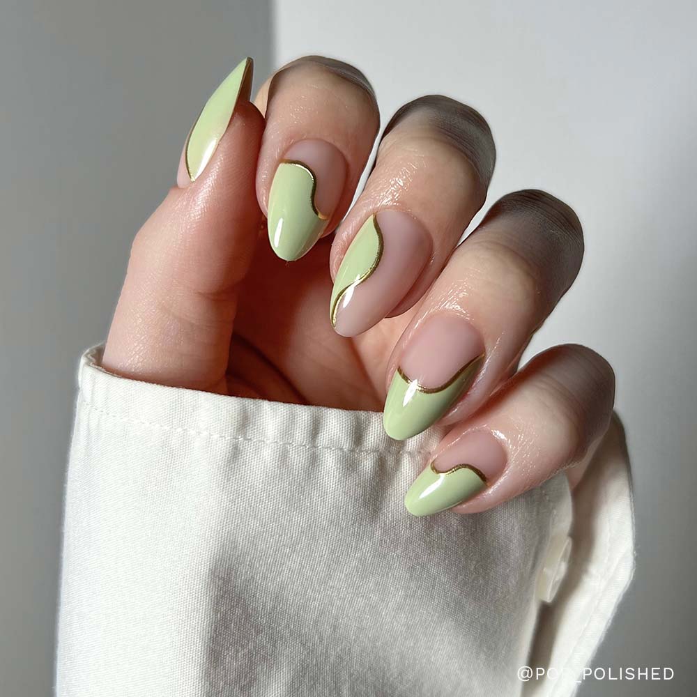 A hand with short almond nails showcases glamnetics Olive You set, featuring pale green and gold abstract wavy French tips. The vegan, cruelty-free nails are shown against a white background, worn by someone in a white long-sleeved shirt.