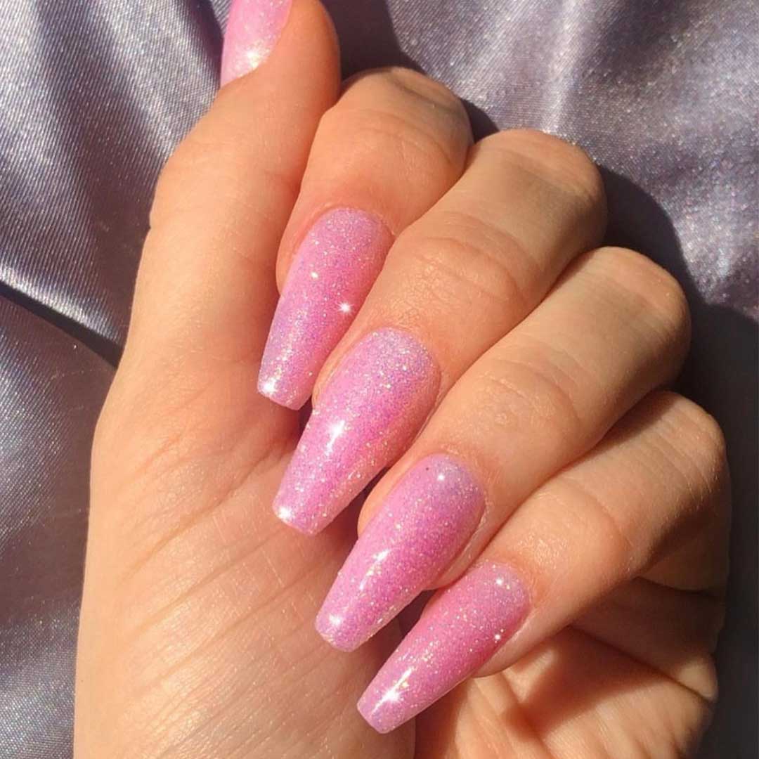 A hand with long, coffin-shaped nails painted in glamnetics Juicy shimmery pink polish reflects light, while a shiny lavender fabric background adds early 2000s vibes.